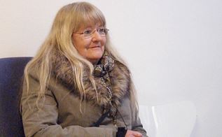 59-årige Joan Thomsen sidder i en stol. Hun har langt hår, briller og frakke på